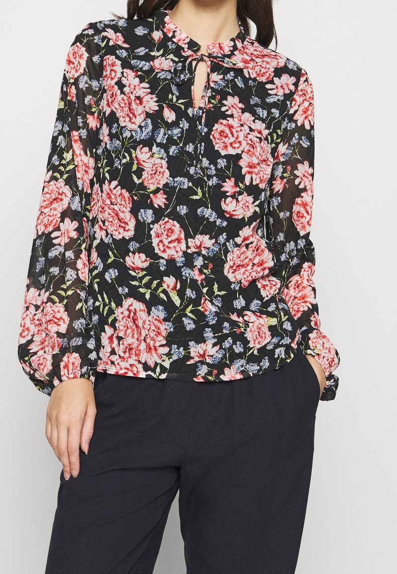 Blouse fleurie en tissu noir transparent avec des fleurs rouges et roses, un col en trou de serrure et des manches longues bouffantes. Associée à un pantalon foncé.