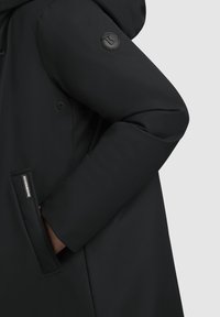 Abrigo acolchado negro con capucha redondeada, un logotipo circular en la manga y un bolsillo lateral con una etiqueta. El material parece suave y mate.