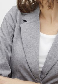 Grå blazer med en strukturerad yta, med en bred krage och en lös passform, lagd över en vit topp. Nära bild av tygdetaljer.