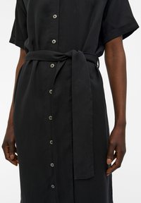 Schwarzes Hemdkleid mit kurzen Ärmeln, einem Gürtel in der Taille und einem geraden Schnitt mit sichtbaren Knöpfen an der Vorderseite.