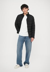 Jachetă puffer neagră cu cusături orizontale, închidere cu fermoar și guler înalt. Purtați-o peste un tricou alb și asortați-o cu blugi de culoare albastră deschisă și adidași.