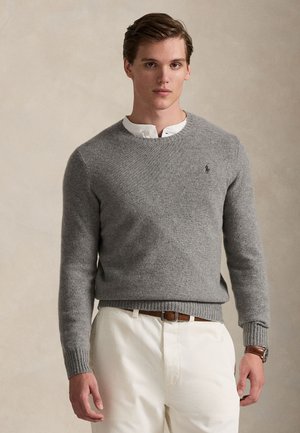 Un mannequin porte un pull gris à col rond par-dessus une chemise blanche et un pantalon crème, sur un fond beige uni.