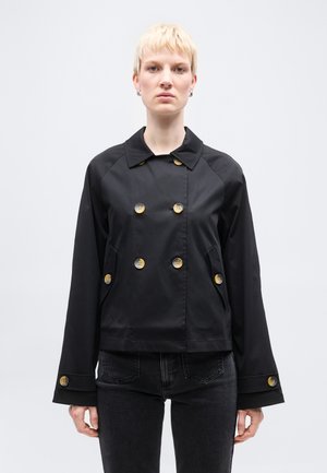 Osoba o krótkich blond włosach, ubrana w czarną, dwurzędową kurtkę z dużymi guzikami i czarne dżinsy, stojąca na tle jednolitego tła.