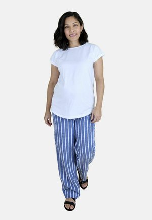 Femeie cu păr castaniu până la umeri, purtând un tricou alb, pantaloni cu dungi albastre și albe și sandale negre, mergând înainte.