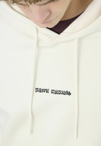 Hoodie couleur crème avec une coupe décontractée, arborant un texte brodé noir "KEITH HARING" et des cordons de serrage. Le matériau semble doux et texturé.