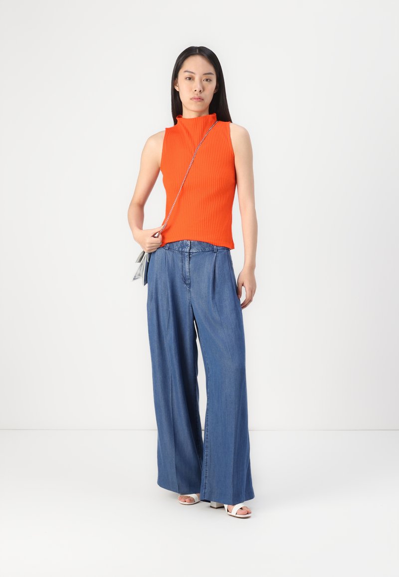 Haut sans manches orange à côtes avec col montant, associé à un pantalon en denim bleu à jambes larges. Le mannequin tient un petit sac argenté et porte des sandales blanches.