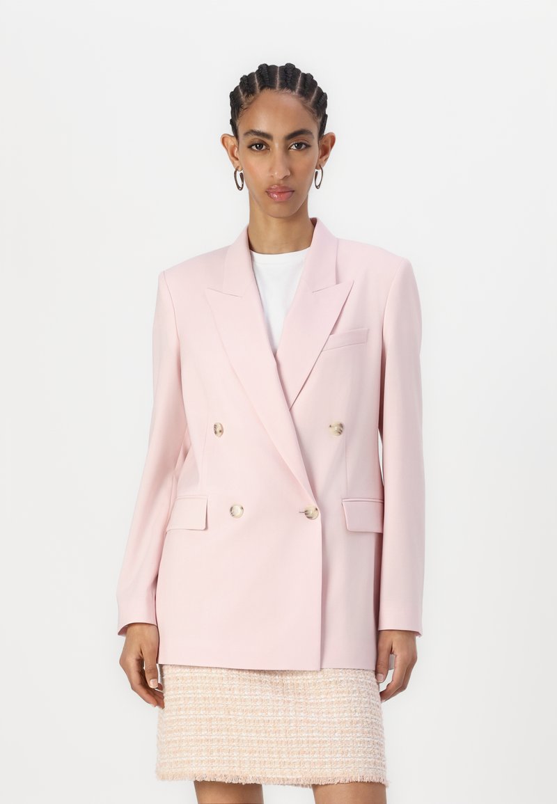 Světle růžový dvouřadý blazer se zlatými knoflíky, s klopami a dvěma předními kapsami, nošený přes bílý top a texturovanou sukni.