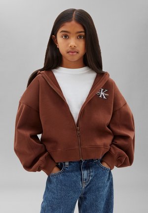 Fiatal személy, hosszú sötét hajjal, barna cipzáras kapucnis pulóvert visel fehér póló és kék farmer felett, kezek zsebbe téve, semleges háttér.