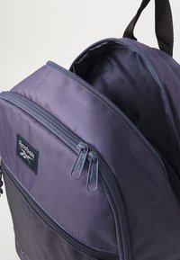 Violet audinių kuprinė su dviem užtrauktukais, logotipo aplikacija ir lygiu viduje. Faktūruota išorė su struktūrine forma ir funkcišku dizainu.
