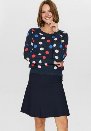 Marineblauer Pullover mit mehrfarbigen Punkten, langen Ärmeln und gewelltem Saum; kombiniert mit einem schwarzen ausgestellten Rock aus glattem Material.
