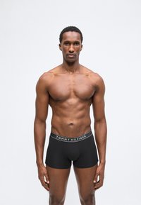 Homme athlétique à la peau foncée debout, portant un boxer noir Tommy Hilfiger, faisant face à l'avant sur un fond blanc uni.