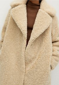 Vaalea, pörröinen, teddy-tyylinen takki, jossa on leveä kaulus ja piilotettu nappikiinnitys. Vaatetuksena ruskea korkeakauluksinen paita. Pehmeä rakenne.