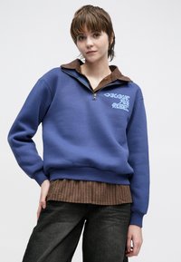 Zils vilnas pulovers ar rāvējslēdzēja apkakli un brūnu imitētu apkakli zem tā. Rotāts ar zilas krāsas tekstu un ribveida aprocēm, valkāts kopā ar brūnu svītrainu kreklu.