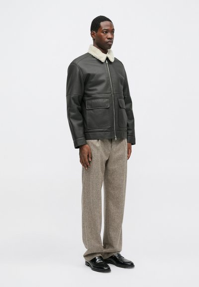Čierna kožená bunda s béžovým ovčím golierom, s dvoma prednými vreckami, skombinovaná so šedými textúrovanými nohavicami a čiernymi topánkami.