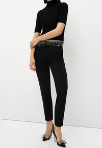 Vrouw in een zwarte korte mouw coltrui met kristalfranje, strakke zwarte broek en zwarte hoge hakken met kristalstrikjes.