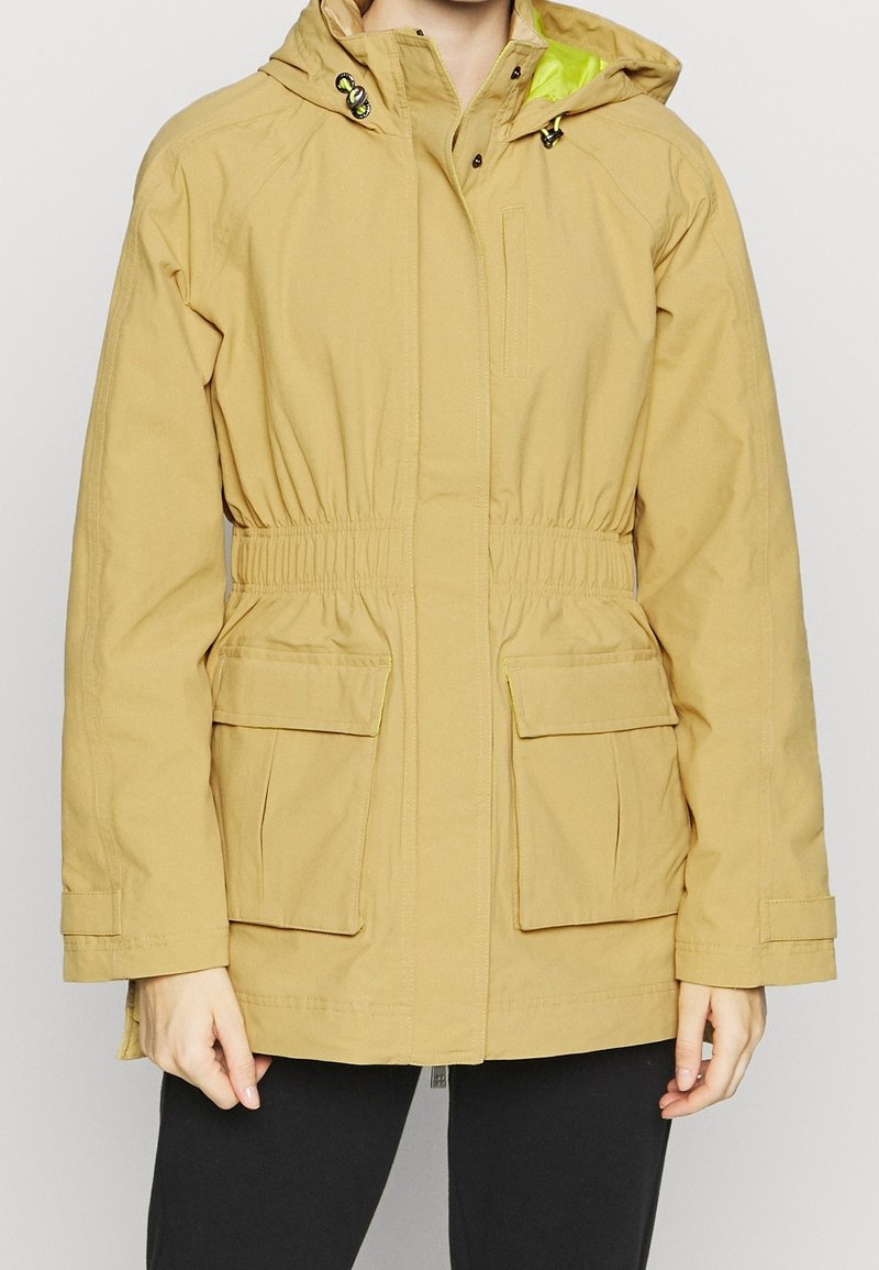 Veste imperméable beige avec une taille cintrée, de grandes poches et une capuche bicolore ornée d'accent jaunes. Texture lisse avec un design contemporain.