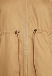 Béžová tkanina se středním zipem a nastavitelnými stahovacími šňůrkami s kovovými zarážkami v oblasti pasu oděvu.