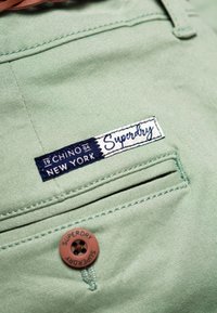 Pantalones chinos verdes claros con una etiqueta azul que lleva el texto blanco "Superdry" y un botón marrón con "Superdry" grabado.