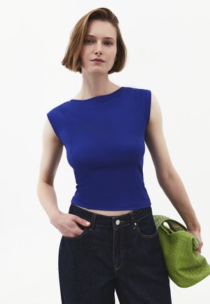 Mujer con cabello corto que lleva una camiseta azul sin mangas y vaqueros oscuros, sosteniendo un bolso tejido verde en la mano izquierda.