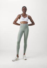 Reggiseno sportivo bianco con spalline regolabili abbinato a leggings alti in un verde chiaro. La modella indossa scarpe da ginnastica bianche. Design semplice eaffusolato.