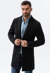 Schwarzer Mantel mit Reverskragen, weicher Textur, einreihigen Knöpfen und seitlichen Taschen, über einem hellblauen Hemd getragen.