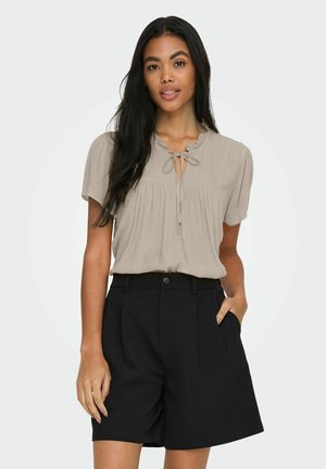 Vrouw draagt een beige blouse met korte mouwen en een strikdetail bij de hals, en zwarte shorts met hoge taille en zakken, staand met één hand in de zak.