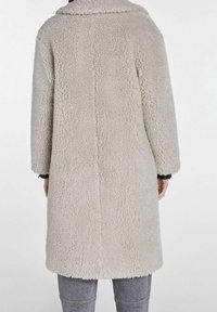 Vue de dos d'une personne portant un long manteau teddy texturé beige clair et un pantalon gris, sur un fond blanc uni.