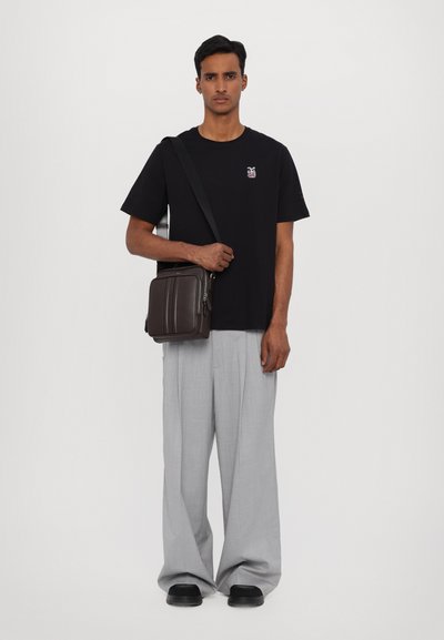 T-shirt noir avec un petit design brodé, pantalon ample gris et un sac bandoulière en cuir marron avec fermeture éclair, se tenant sur un fond uni.