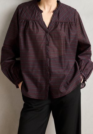 Femme portant un chemisier rayé bordeaux et gris à boutons, manches longues, et un pantalon noir, mains dans les poches.