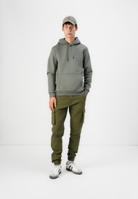 Green hoodie with a front pocket, paired with olive cargo pants featuring side pockets. Completed with a light-cap and white sneakers with black accents.