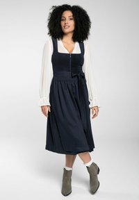 Marine jurk met een getailleerde lichaamsvoering, ritssluiting en een uitlopende rok. Witte blouse met lange mouwen eronder. Grijze enkellaarzen met een chunky hak.