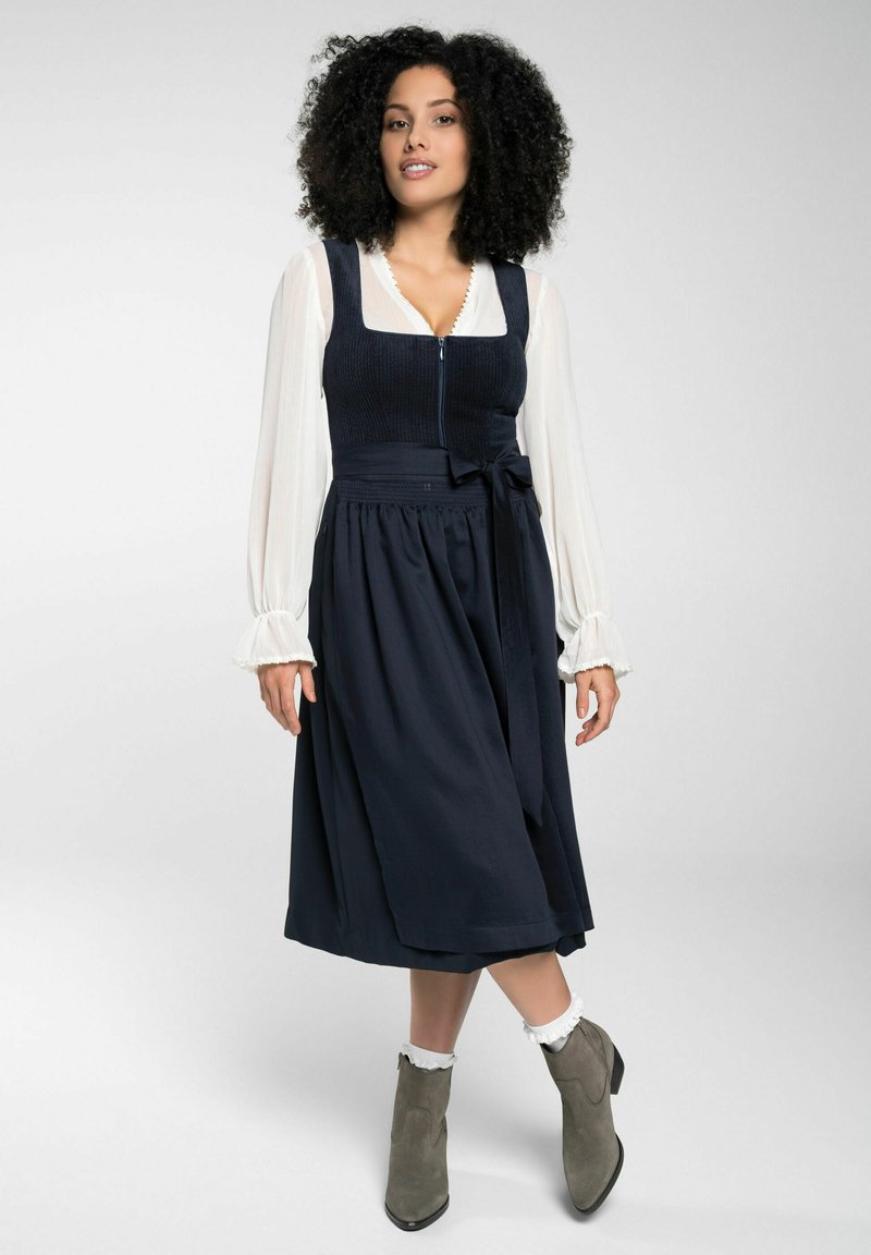 Marine jurk met een getailleerde lichaamsvoering, ritssluiting en een uitlopende rok. Witte blouse met lange mouwen eronder. Grijze enkellaarzen met een chunky hak.