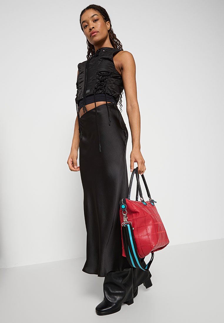 Mujer vistiendo una blusa sin mangas negra y una falda larga, sosteniendo un bolso rojo con una correa a rayas azules, de pie contra un fondo blanco.