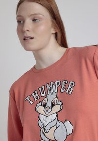 Joven con cabello rojo que lleva un sudadera color coral con el conejo de dibujos animados Thumper y un fondo blanco.