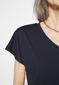 Laivastonsininen V-painikkeinen T-paita, jossa on lyhyet hihat ja pehmeä, sileä kangas. Huomionarvoisia yksityiskohtia ovat huomaamaton ompelu hihansuiden reunalla.