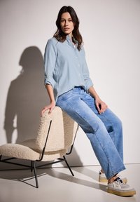 Camicia a righe blu chiaro con bottoni, vestibilità rilassata, abbinata a jeans blu chiaro a gamba dritta. Seduto su una sedia beige, con texture, e gambe nere.