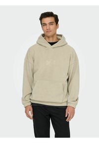 Beżowa bluza polarowa z dużą przednią kieszenią, regulowanym kapturem ze sznurkiem i prążkowanymi mankietami, noszona na czarnej koszulce, w połączeniu z czarnymi jeansami.