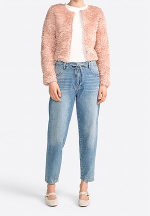 Kvinde iført en lyserød fluffy cropped jakke, hvid top, lyseblå højtaljede jeans med snor og cremefarvede Mary Jane-sko.