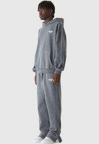 Szürke, túlméretezett kapucnis pulóver kenguru zsebbel és hímzett logóval, amelyhez oldalsávos, elasztikus mandzsettás derékig érő nadrág illik.