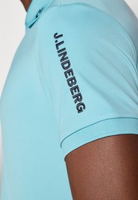 Lys blå polo skjorte laget av glatt stoff. Har en ribbet krage og svart brodert logo på ermet.