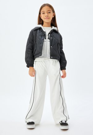 Dziewczyna stojąca, ubrana w czarną pikowaną kurtkę, szarą bluzę z kapturem, szerokie białe spodnie z czarnymi pasami i czarne trampki na białym tle.