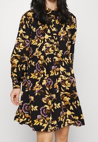 Mujer vestida con un vestido negro de manga larga con patrones florales en amarillo, morado y marrón, con un cuello y botones en la parte delantera.