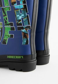 Siniset kumisaappaat, joissa on mustia yksityiskohtia, tekstuuripohjat, rohkea "MINECRAFT" -merkintä vihreänä ja värikkäitä pikselöityjä suunnitteluelementtejä.