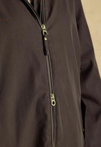 Tuva tuvumā redzams tumši brūns jakas ar diviem zelta rāvējslēdzējiem, viens daļēji atvērts augšpusē, bet otrs pilnībā aizvērts vidusdaļā.