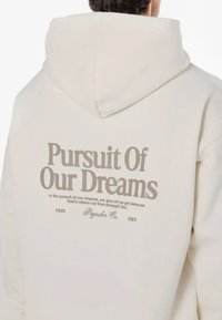 Sudadera de color crema con un gran estampado gráfico en la parte trasera. El texto dice "Búsqueda de Nuestros Sueños" en letras marrones en negrita. Textura de tela suave.