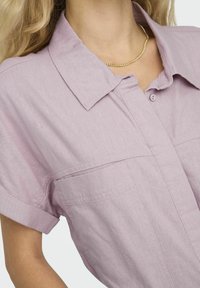 Camisa de botones de color morado claro con cuello, mangas cortas dobladas y bolsillos frontales, hecha de una tela texturizada. Acento de cadena dorada visible.