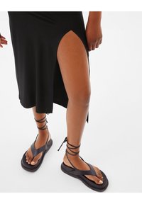 Svart klänning med en hög slits vid låret, matchad med platta svarta sandaler med korsade ankelremmar och en tjock sula.