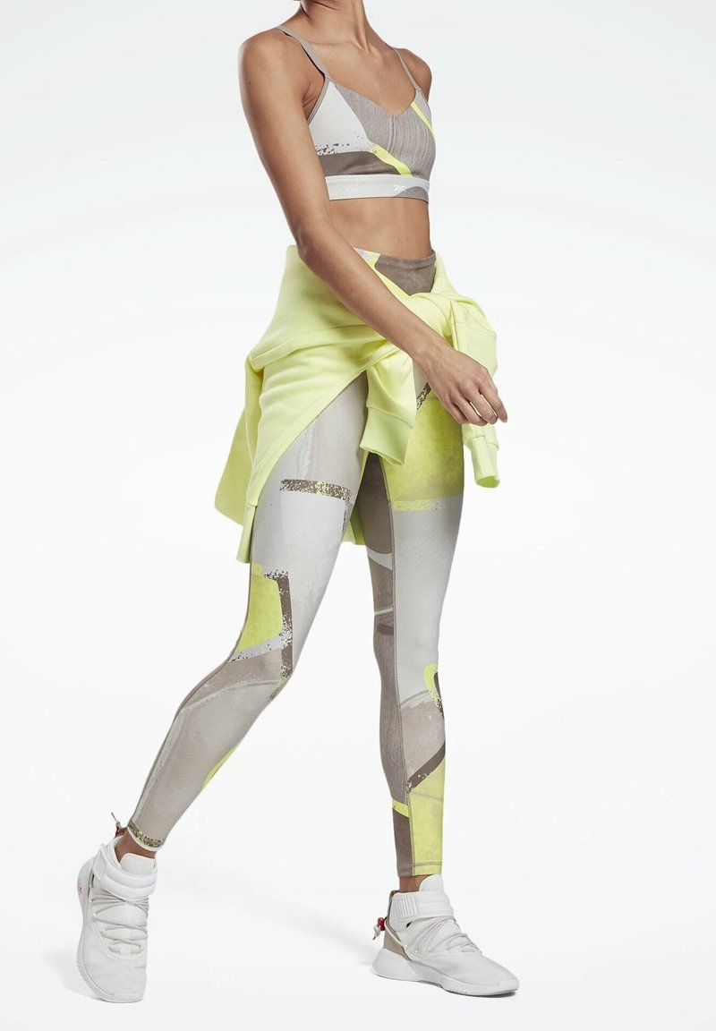 Kvinna som bär ett mönstrat sport-bh och leggings med en knytet gul sweatshirt och vita träningsskor, stående i en gångposition.