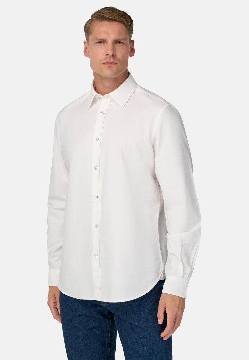 Uomo che indossa una semplice camicia bianca a maniche lunghe e jeans blu, in piedi contro uno sfondo chiaro e neutro.