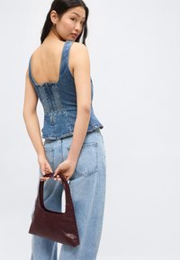 Γυναίκα που φοράει αμάνικο τζιν τοπ και ανοιχτόχρωμα μπλε τζιν παντελόνια, κρατώντας μια μικρή σκούρα καφέ δερμάτινη τσάντα πίσω από την πλάτη της.